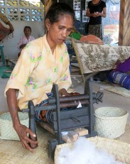 A woman feeds a cotton boll through two rollers in a wooden mangle to soften the fibres. This process also separates the cotton seeds which can then be replanted in communal gardens.