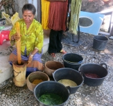 A woman pounds turmeric bulbs and water to make a natural yellow dye. Behind her hang skeins of naturally-died cotton.